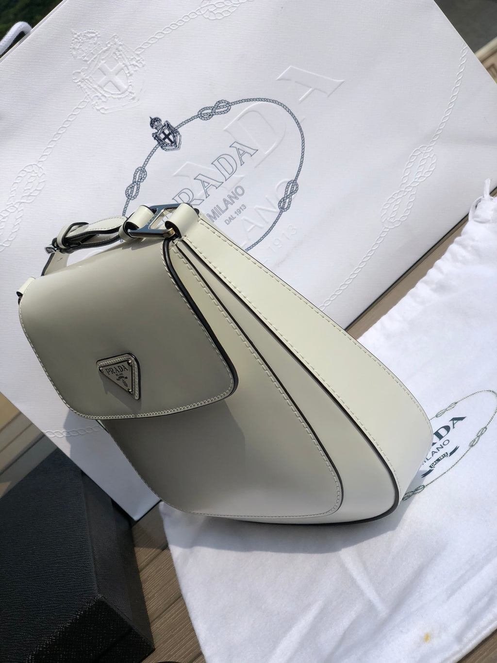 白色❗️尺寸：19*6*24cm（56980898）PRADA代工廠出品翻蓋腋下包❗️普拉達這款精緻的Prada Cleo手袋重新詮釋品牌標誌性的復古設計。底部和側面獨特的傾斜結構彰顯這款翻蓋手袋簡約的弧形線條，賦予其輕盈柔和的造型風格。 Prada系列別緻的亮面皮革材質頗具現代感與多變風格，慣於創造嶄新的組合與對比效果。琺瑯三角形標誌呈現鮮明的品牌格調。磁扣扣合Prada標誌尼龍襯裡，配上貼布口袋。可延長皮革提把，可將單肩包變成斜背包！