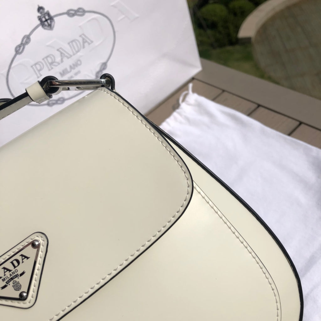 白色❗️尺寸：19*6*24cm（56980898）PRADA代工廠出品翻蓋腋下包❗️普拉達這款精緻的Prada Cleo手袋重新詮釋品牌標誌性的復古設計。底部和側面獨特的傾斜結構彰顯這款翻蓋手袋簡約的弧形線條，賦予其輕盈柔和的造型風格。 Prada系列別緻的亮面皮革材質頗具現代感與多變風格，慣於創造嶄新的組合與對比效果。琺瑯三角形標誌呈現鮮明的品牌格調。磁扣扣合Prada標誌尼龍襯裡，配上貼布口袋。可延長皮革提把，可將單肩包變成斜背包！