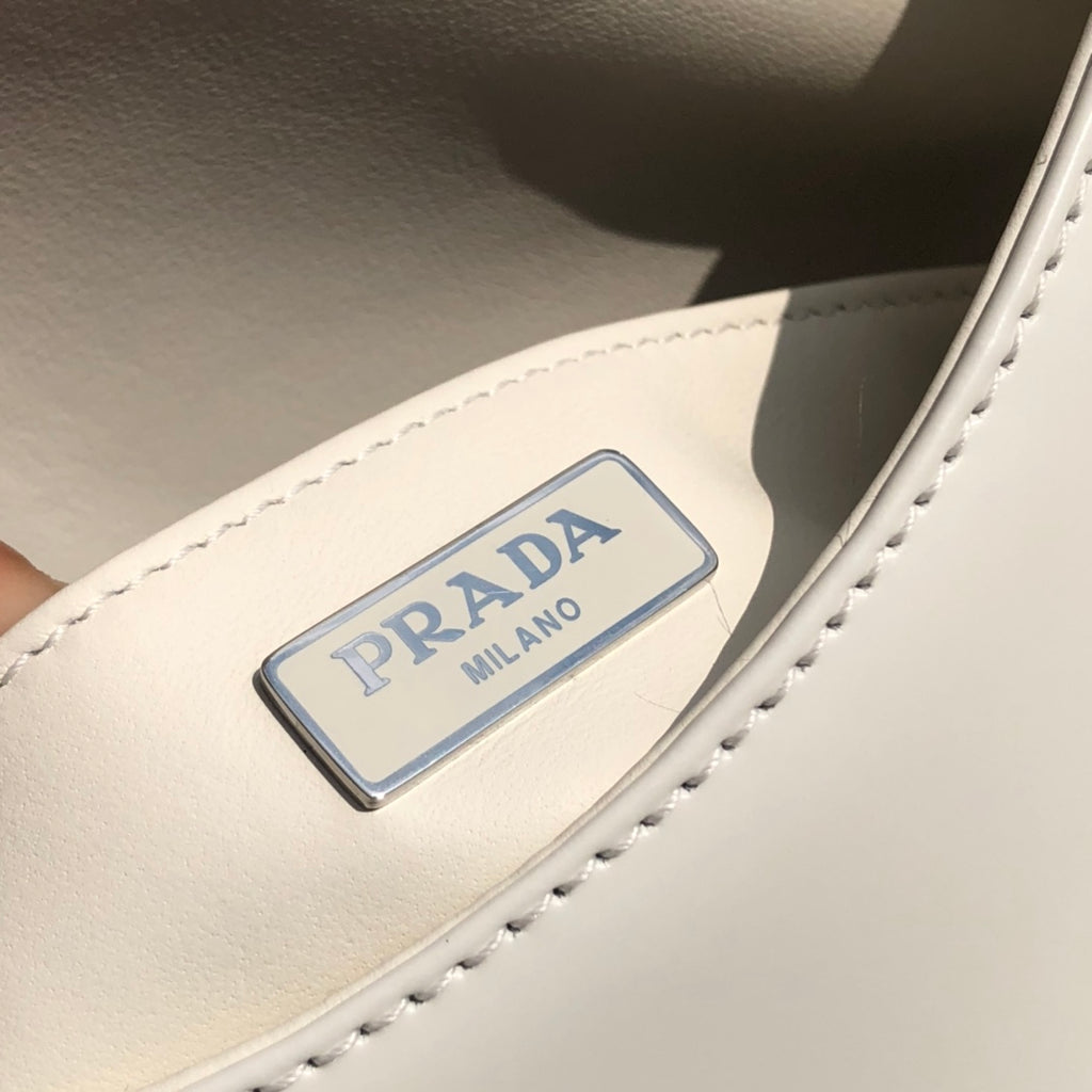 白色❗️尺寸：19*6*24cm（56980898）PRADA代工廠出品翻蓋腋下包❗️普拉達這款精緻的Prada Cleo手袋重新詮釋品牌標誌性的復古設計。底部和側面獨特的傾斜結構彰顯這款翻蓋手袋簡約的弧形線條，賦予其輕盈柔和的造型風格。 Prada系列別緻的亮面皮革材質頗具現代感與多變風格，慣於創造嶄新的組合與對比效果。琺瑯三角形標誌呈現鮮明的品牌格調。磁扣扣合Prada標誌尼龍襯裡，配上貼布口袋。可延長皮革提把，可將單肩包變成斜背包！