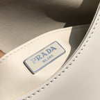 白色❗️尺寸：19*6*24cm（56980898）PRADA代工廠出品翻蓋腋下包❗️普拉達這款精緻的Prada Cleo手袋重新詮釋品牌標誌性的復古設計。底部和側面獨特的傾斜結構彰顯這款翻蓋手袋簡約的弧形線條，賦予其輕盈柔和的造型風格。 Prada系列別緻的亮面皮革材質頗具現代感與多變風格，慣於創造嶄新的組合與對比效果。琺瑯三角形標誌呈現鮮明的品牌格調。磁扣扣合Prada標誌尼龍襯裡，配上貼布口袋。可延長皮革提把，可將單肩包變成斜背包！
